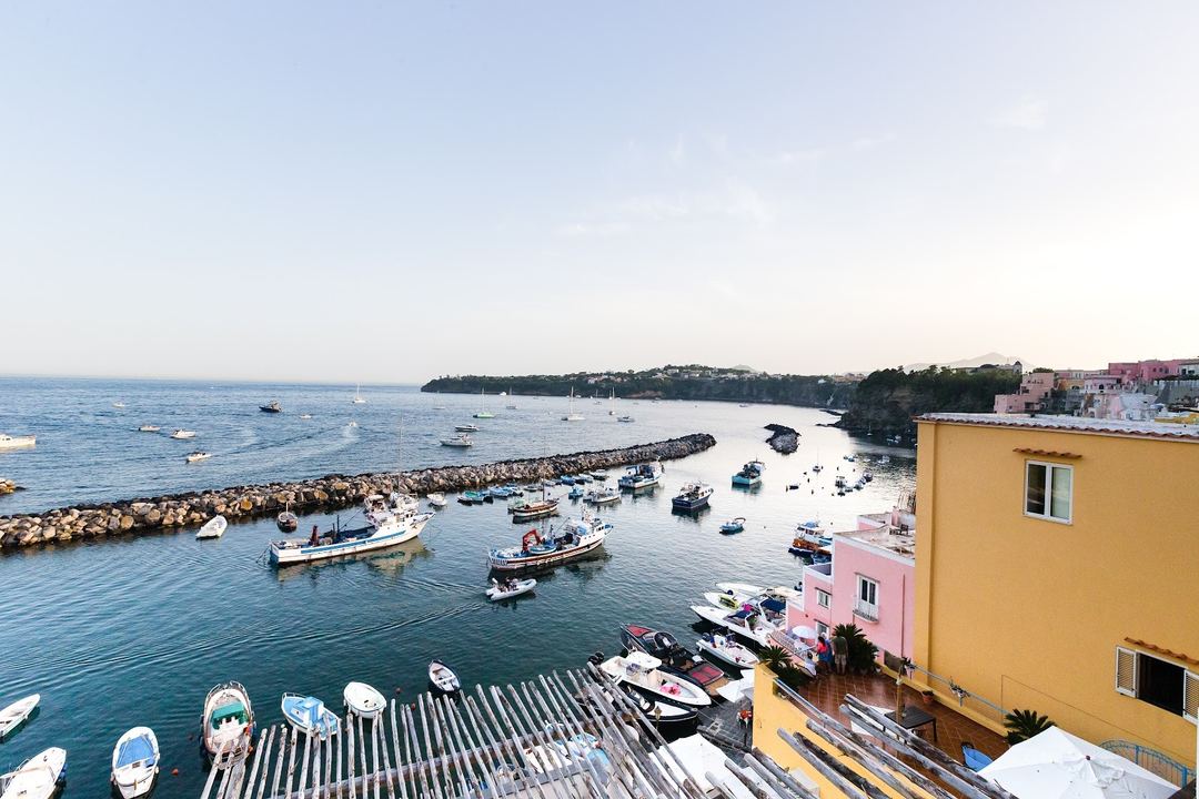 Panoramic view of Procida's Marina Grande harbor with colorful boats, traditional architecture, and rocky breakwater against clear blue Mediterranean waters.