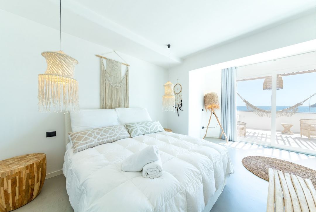 A bright, minimalist bedroom with a white bed, woven pendant lights, and direct access to a sea-view terrace.