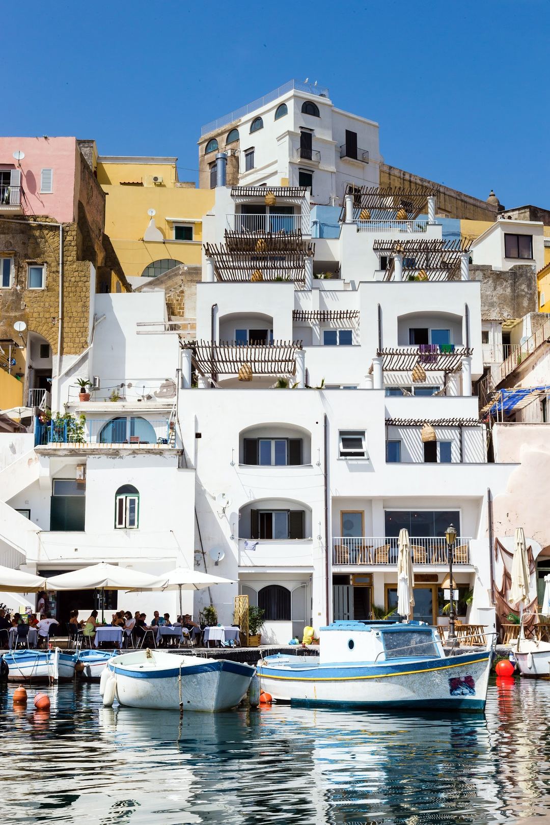 Waterfront view of a white Mediterranean-style hotel building with multiple balconies overlooking the harbor with traditional fishing boats.