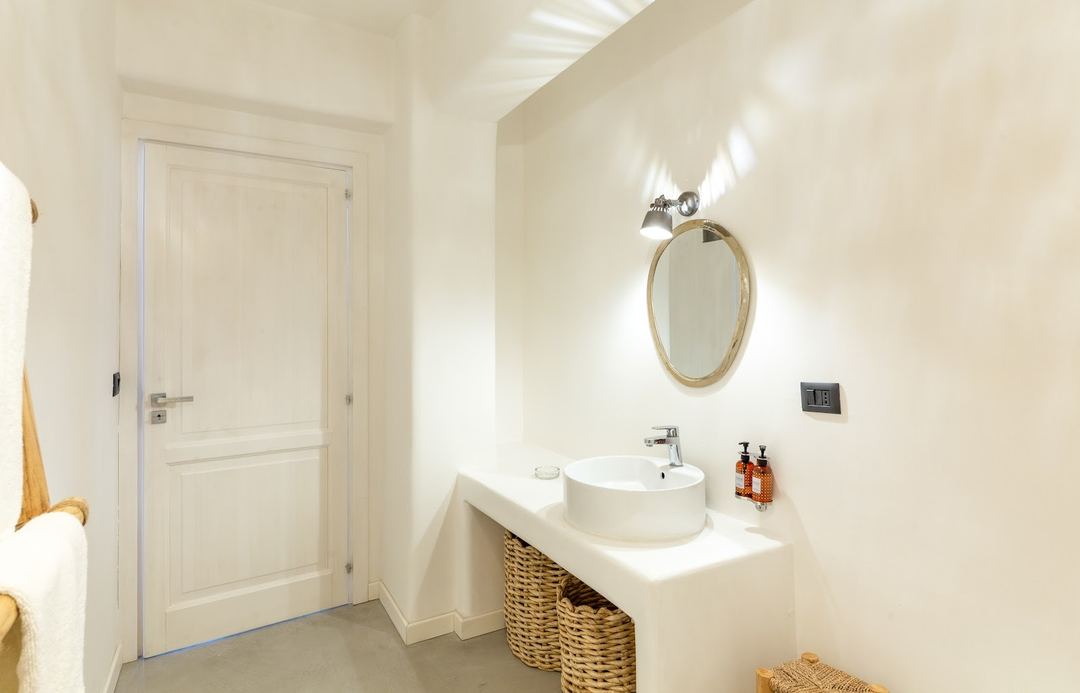 A bright, minimalist bathroom features a round white vessel sink on a woven basket vanity, with an oval mirror and chrome fixtures.