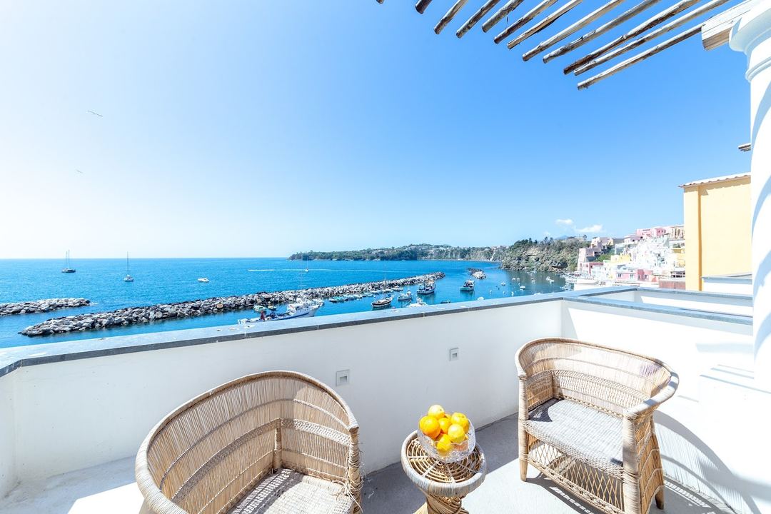 A private terrace with wicker seating overlooks a picturesque coastal harbor with colorful buildings and moored boats in the Mediterranean.