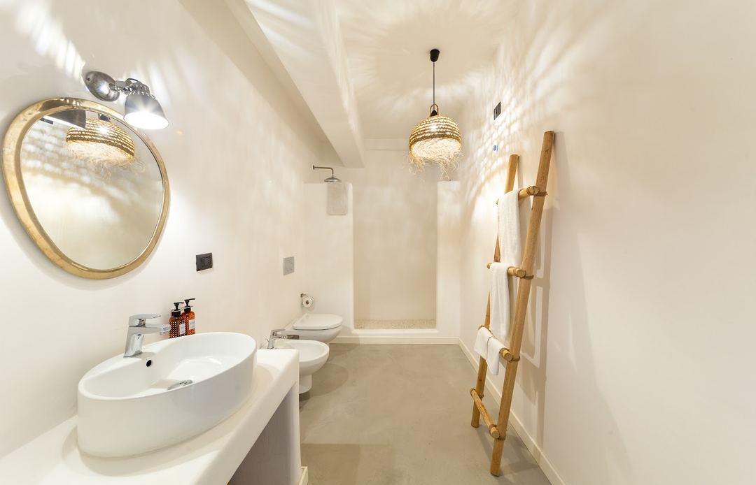 A modern bathroom features a curved white sink, gold-framed mirror, pendant lighting, and a wooden towel ladder against cream-colored walls.