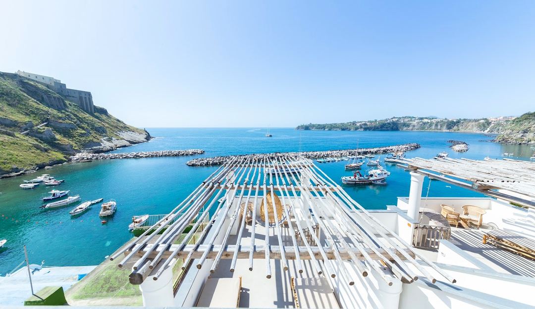 Aerial view of Procida harbor with moored boats, white dock structures, and colorful coastal cliffs under clear blue skies.