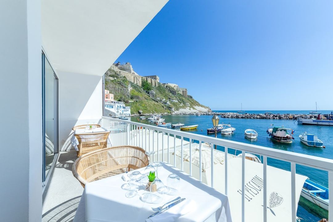 A waterfront balcony dining setup overlooks the harbor of Procida with anchored fishing boats and a historic fortress on the hillside.