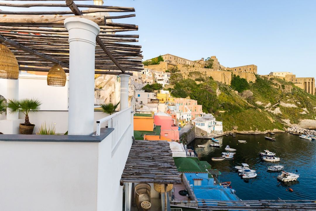 A panoramic view from the hotel's terrace overlooking the colorful harbor of Procida with moored fishing boats and the historic aragonese castle ruins perched on the clifftop.